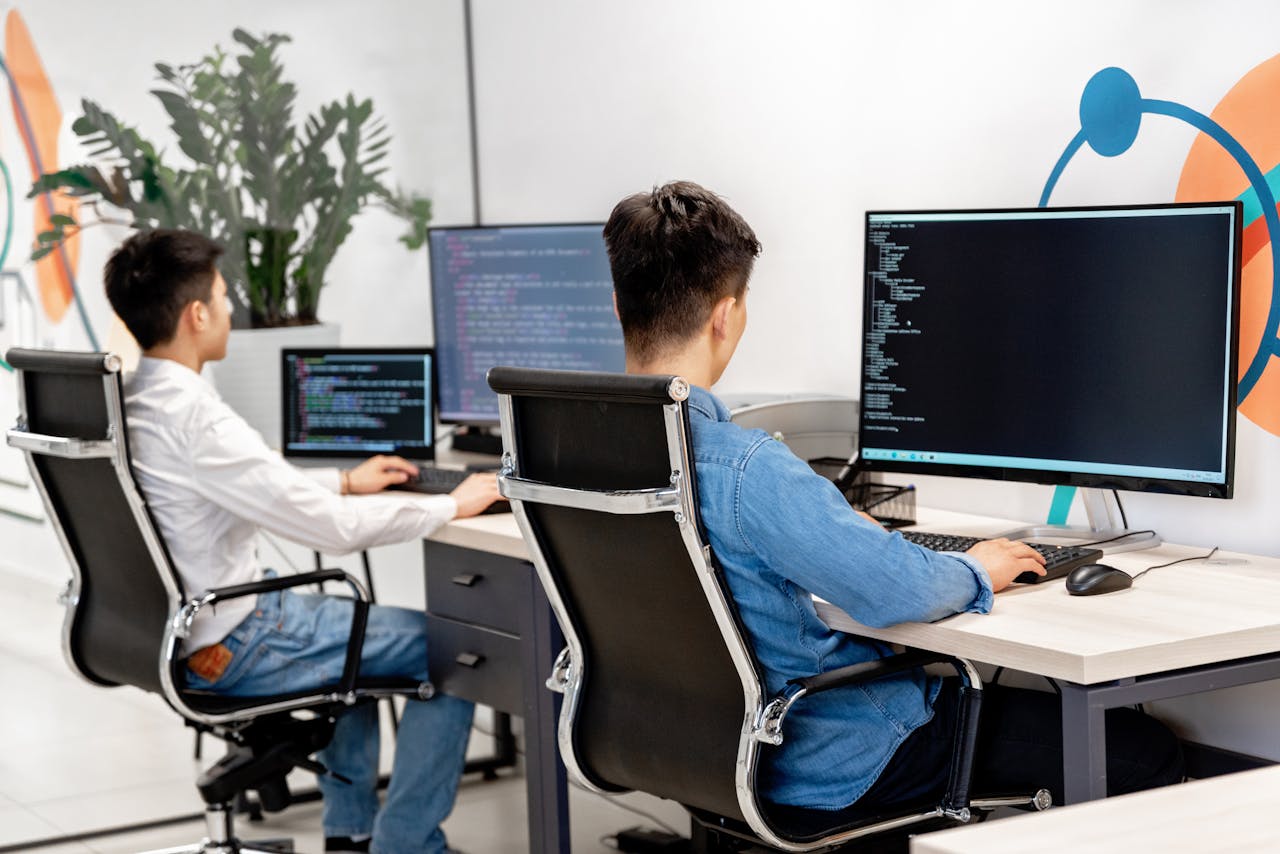 services-04 Two male developers at desks programming in a modern office workspace with large monitors.